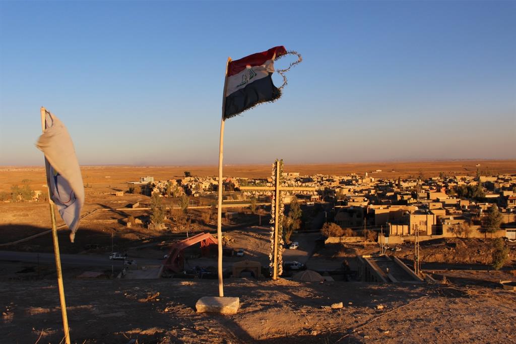 La bandiera delle milizie cristiane e dell'Iraq dietro a una croce sulla collina da cui si vede la pianura di Ninive