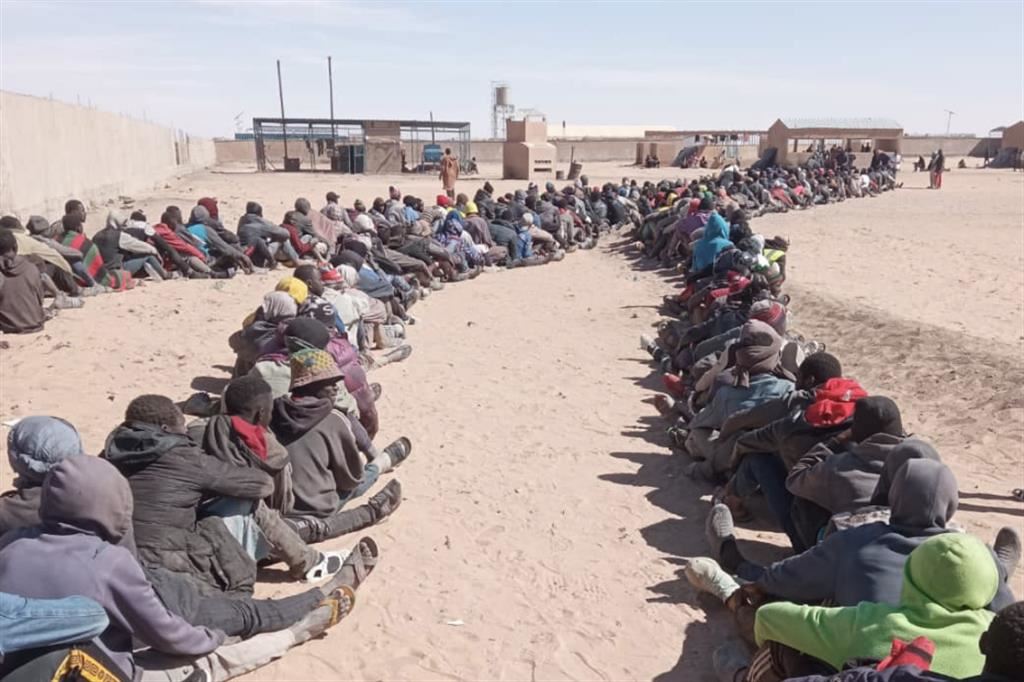 Centinaia di persone sedute, in fila, nel deserto di Assamaka, in Niger